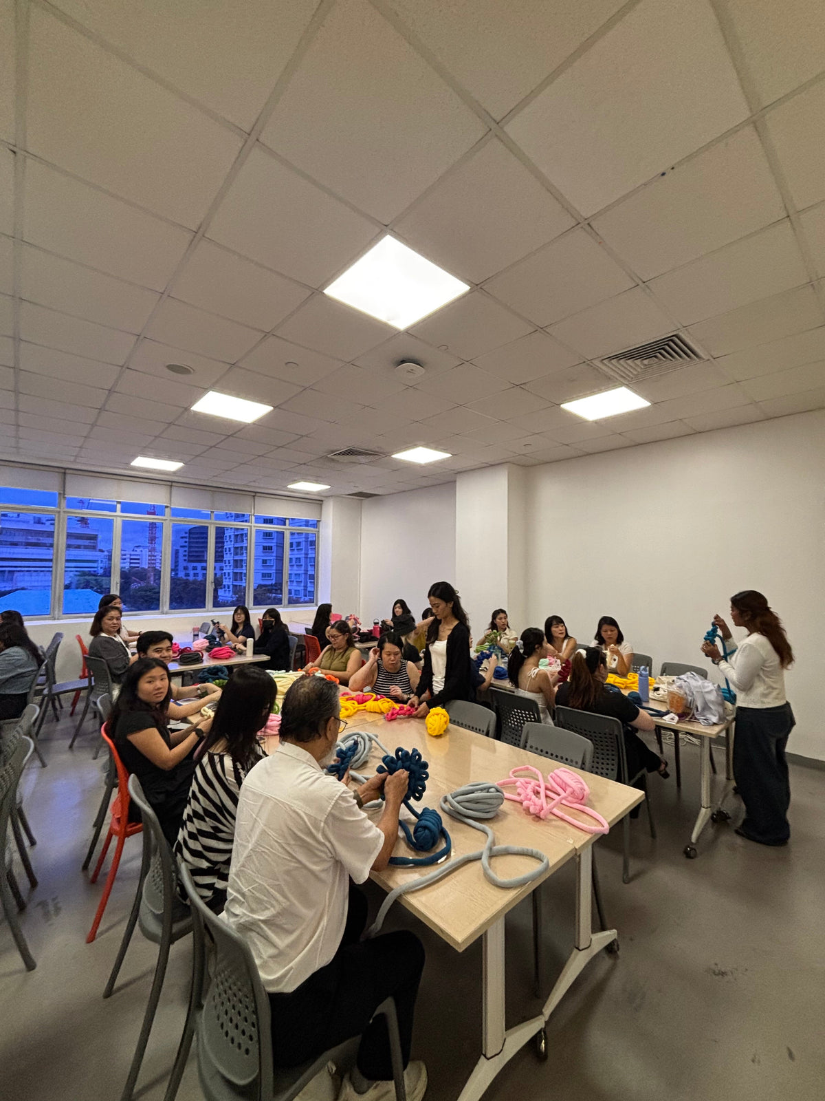 Wide shot of a large group of participants at a NAFA workshop seated around long tables in a classroom setting, hand-weaving colorful chunky yarn bags with skeins of blue, yellow, and pink yarn visible