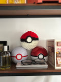 Three crocheted versions of Poké Ball on a shelf with a bottle and glasses.