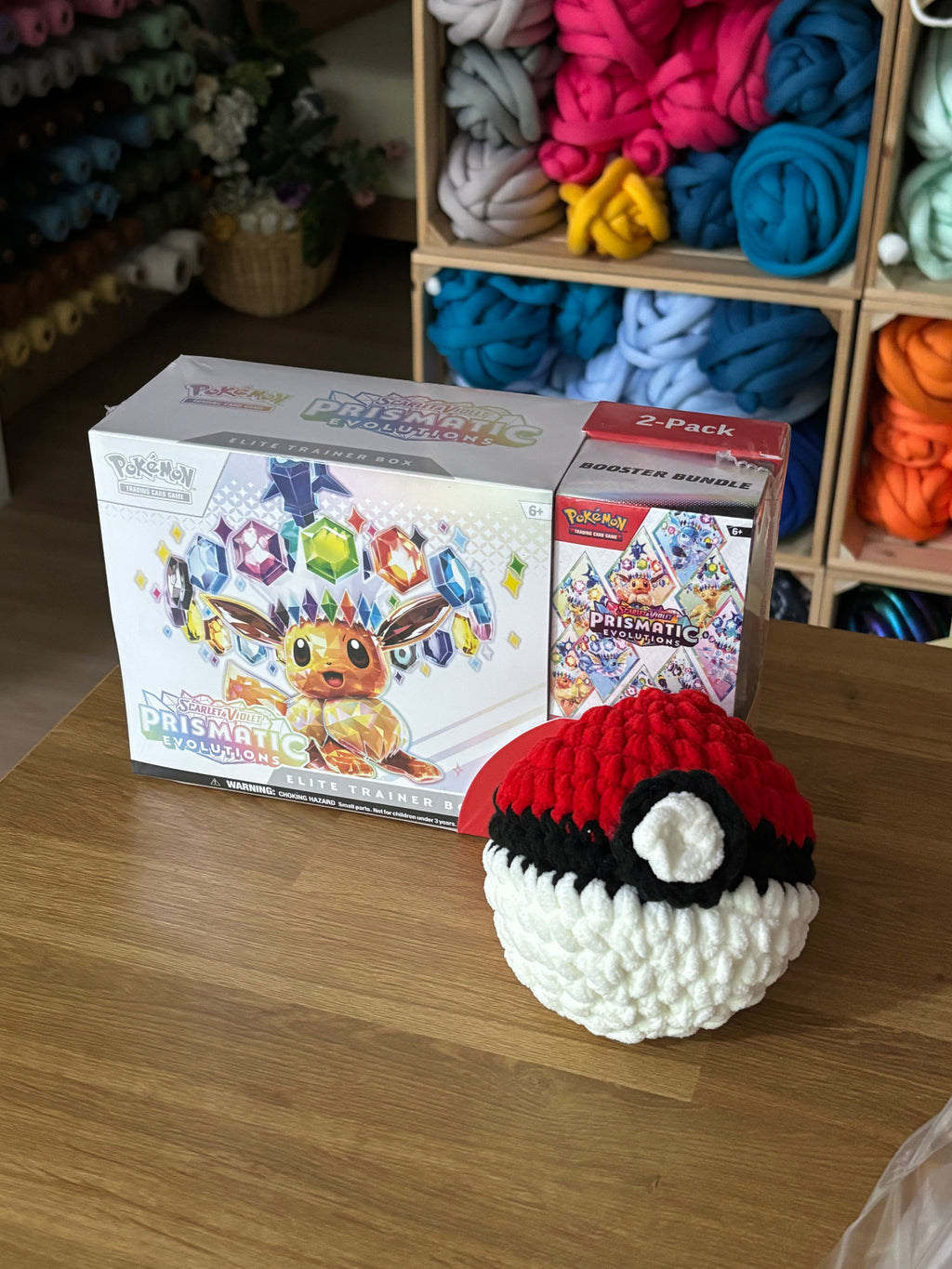 Crocheted Poké Ball on a wooden surface with a box of Poké Ball plush toys in the background.