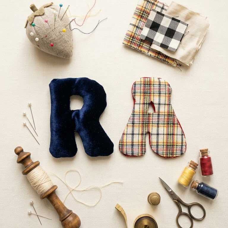 A styled flat-lay of sewing and fabric crafting supplies on a light cream background, including a navy blue velvet letter R, a plaid fabric letter A, a pincushion, folded plaid and cream fabric, thread, scissors, a wooden spool, pins, and small spools of thread.