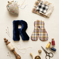 A styled flat-lay of sewing and fabric crafting supplies on a light cream background, including a navy blue velvet letter R, a plaid fabric letter A, a pincushion, folded plaid and cream fabric, thread, scissors, a wooden spool, pins, and small spools of thread.