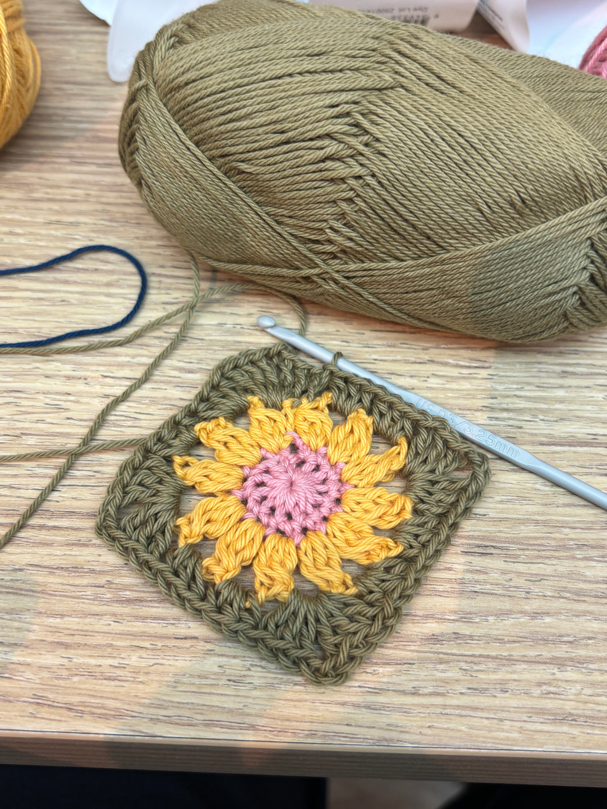 A work-in-progress handmade crochet granny square featuring a textured sunflower motif with a pink center, bright yellow petals, and an olive green border, resting on a wooden table next to a large skein of olive green yarn and a silver crochet hook.