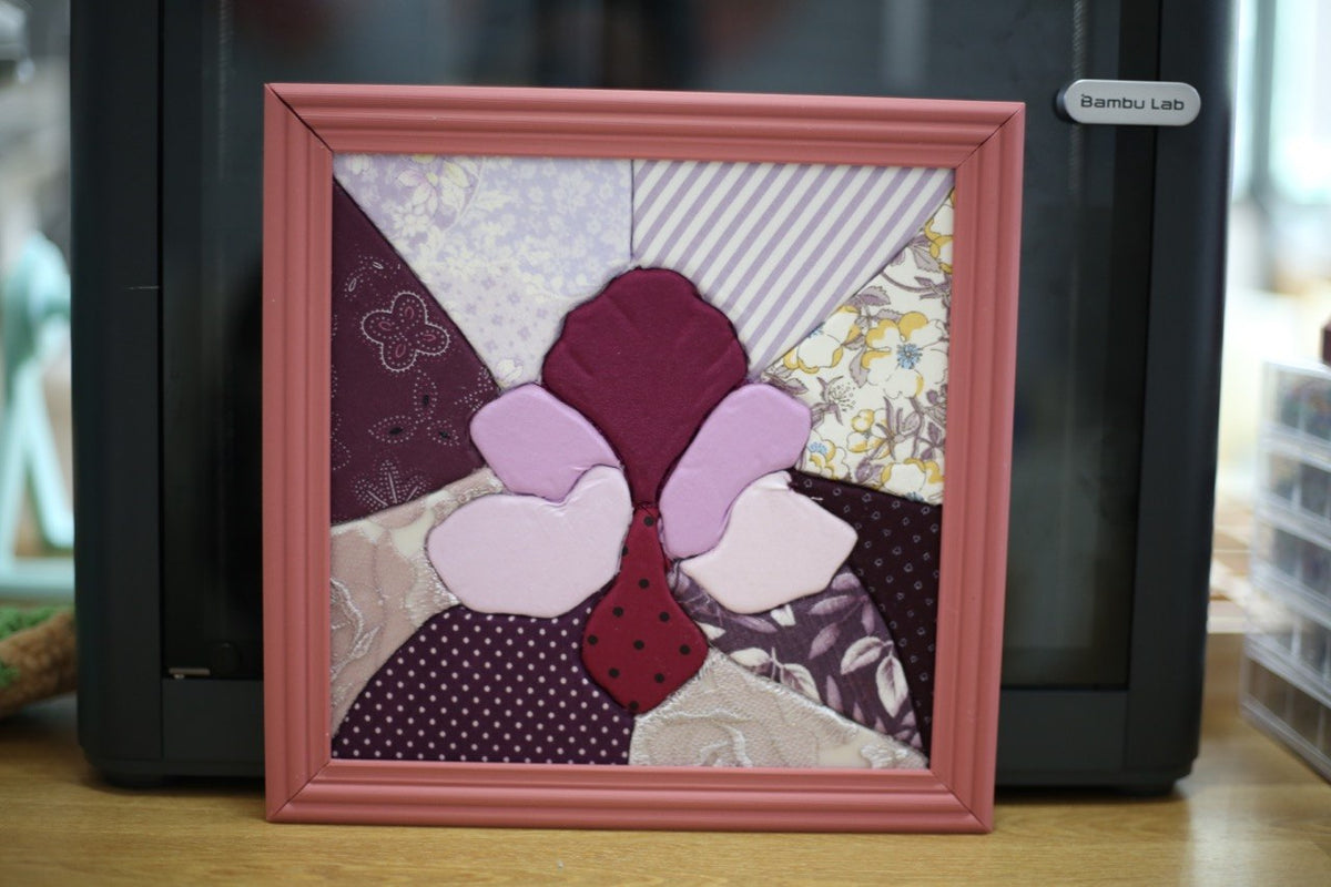 A completed square Kinusaiga fabric art piece featuring a stylized orchid design made from various purple, pink, and patterned textile scraps, enclosed in a dusty pink wooden frame.
